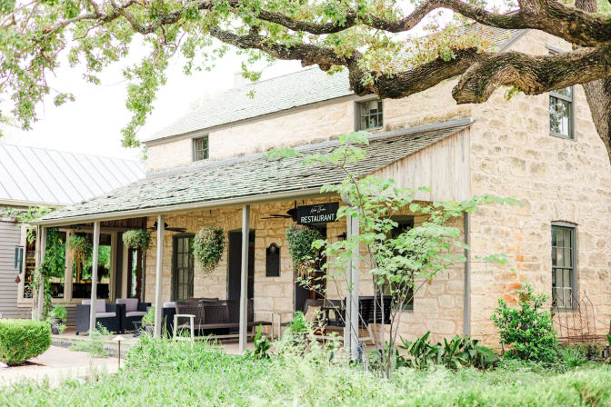 Rustic stone country restaurant with covered porch, hanging planters, outdoor seating and lush garden under large oak tree branches