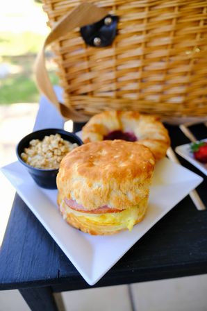 Flaky biscuit breakfast sandwich with egg, cheese and ham on a white plate, small granola cup and jam-filled pastry beside a wicker picnic basket for outdoor brunch