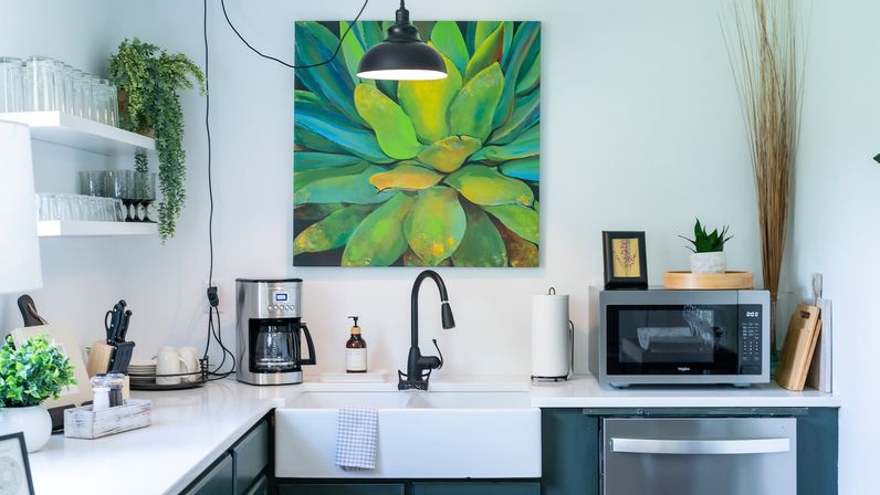 Bright modern home kitchen with a white farmhouse sink and black gooseneck faucet, coffee maker and microwave on a white countertop, large green succulent painting above, potted plants, glassware on open shelves and wooden cutting boards.