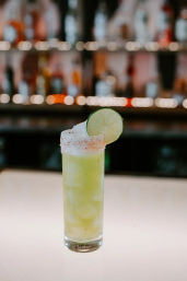 Tall green lime cocktail in a salt-rimmed highball glass with a lime wheel garnish on a backlit bar countertop and blurred bottles with bokeh lights in the background