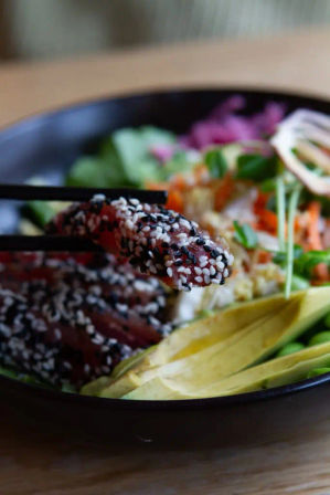 Close-up sesame-crusted tuna poke bowl with sliced avocado, mixed greens, shredded carrots and pickled cabbage, chopsticks holding a sesame-coated tuna slice