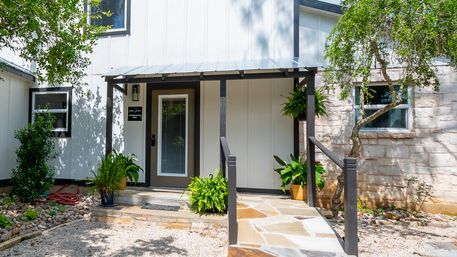 Charming covered front porch of a white farmhouse-style home with black trim, glass-panel door, stone walkway, potted ferns and gravel yard under shaded trees