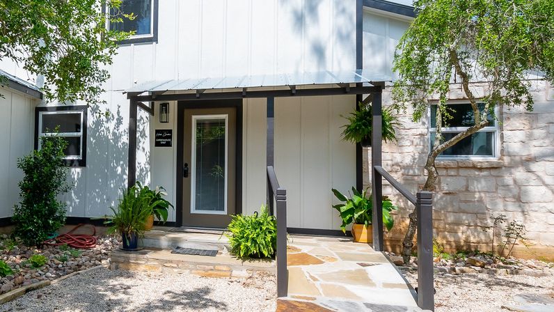 Charming covered front porch of a white farmhouse-style home with black trim, glass-panel door, stone walkway, potted ferns and gravel yard under shaded trees