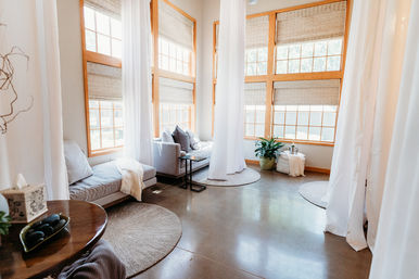 Sunlit modern sitting room with tall wood-framed windows, sheer white curtains, gray sofas, round woven rugs, a potted plant, and polished concrete floor.