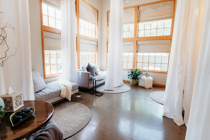 Sunlit modern sitting room with tall wood-framed windows, sheer white curtains, gray sofas, round woven rugs, a potted plant, and polished concrete floor.