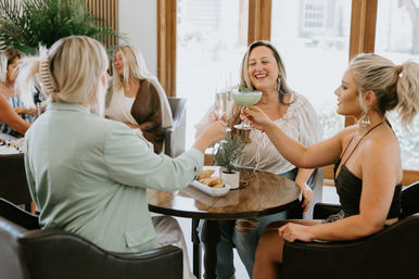 Three women laughing and clinking cocktails at a sunlit cafe brunch table with pastries and a small potted plant