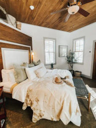 Cozy modern farmhouse bedroom interior with wood-paneled ceiling and fan, white rumpled bedding topped with a straw hat, woven bench and potted plant by sunny windows