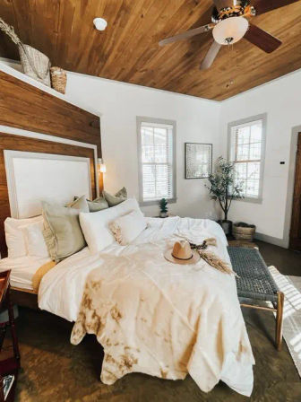 Cozy modern farmhouse bedroom interior with wood-paneled ceiling and fan, white rumpled bedding topped with a straw hat, woven bench and potted plant by sunny windows