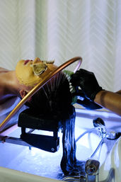 Client reclined at a salon sink having long dark hair rinsed under a copper shower ring by a gloved stylist; golden facial mask with tea bags over eyes and blue-lit basin.