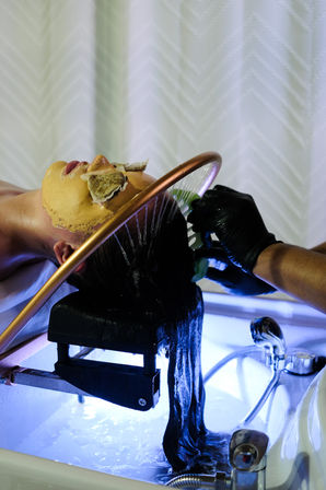 Client reclined at a salon sink having long dark hair rinsed under a copper shower ring by a gloved stylist; golden facial mask with tea bags over eyes and blue-lit basin.