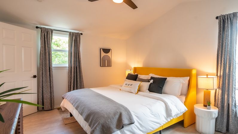 Bright modern bedroom with a yellow upholstered bed, white bedding and gray throw, matching gray curtains, wood floor, bedside lamps and minimalist wall art — cozy contemporary guest room.