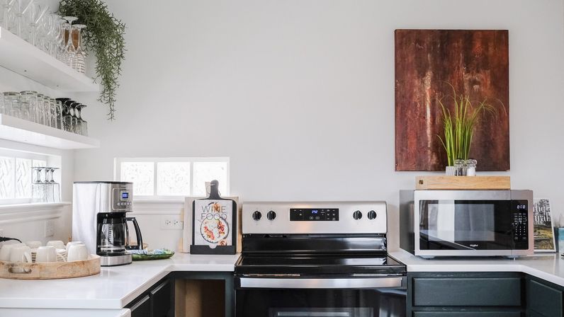 Sunlit modern kitchen corner with stainless steel electric range and microwave, coffee maker on white countertops, open shelves of glassware, a wooden tray of mugs, potted greenery and rust-colored wall art.
