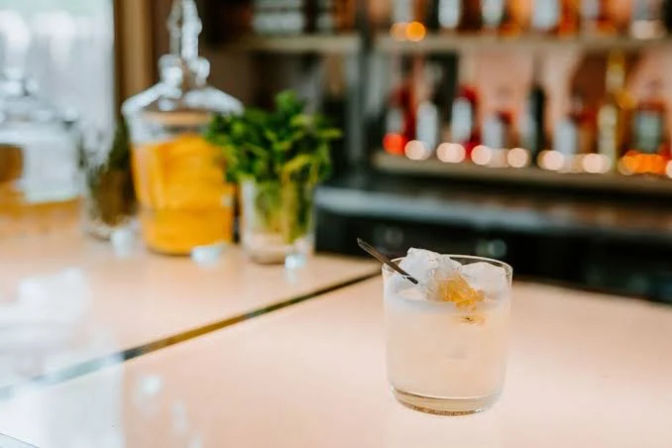 Craft cocktail in a short glass with crushed ice and citrus peel on a glowing bar counter, herb jar and blurred liquor bottles in the background