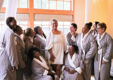 Laughing multi-ethnic group of women in matching gray robes gathered around a woman in a white dress in a sunlit dressing room by wooden windows — relaxed, joyful getting-ready moment.