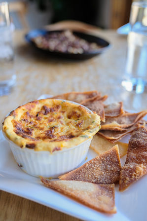 Melty baked cheese dip in a white ramekin served with golden crispy chips on a white plate on a wooden table — casual restaurant appetizer.