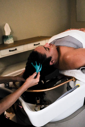 Client reclined at a salon shampoo bowl receiving a scalp massage with a teal silicone massager as stylist washes long curly hair
