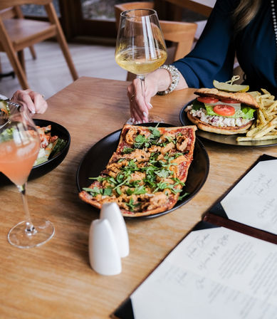 Casual bistro table with rectangular flatbread topped with arugula and fried onions, a sandwich with lettuce and tomato and fries, a glass of white wine, a pink cocktail and visible menus — brunch or lunch scene.