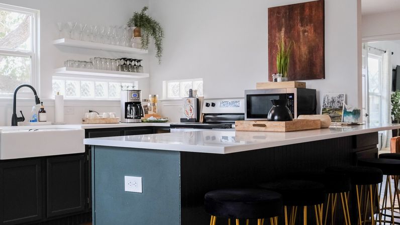 Bright modern kitchen with white quartz island and dark cabinets, farmhouse sink, open glass shelves with glassware, coffee maker, potted plant, and black bar stools.
