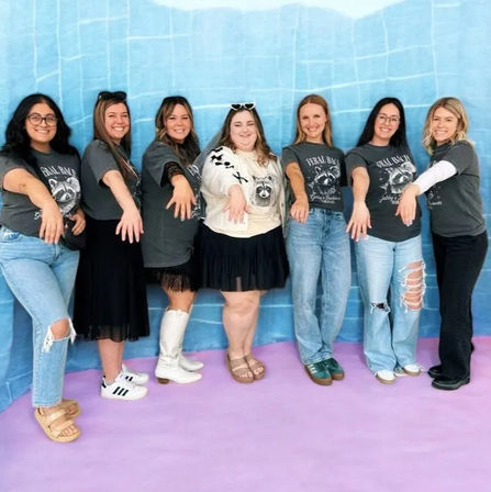 Seven women in casual denim and skirts posing in front of a blue backdrop and pink floor, wearing matching dark graphic tees and extending hands forward to show rings in a fun group portrait.