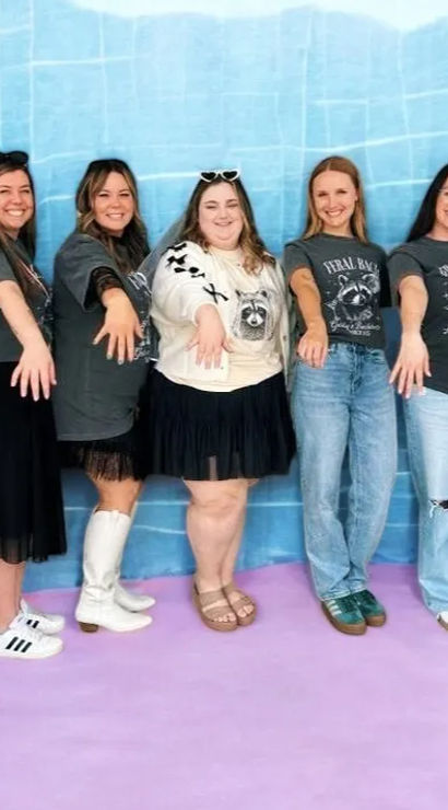 Seven women in casual denim and skirts posing in front of a blue backdrop and pink floor, wearing matching dark graphic tees and extending hands forward to show rings in a fun group portrait.