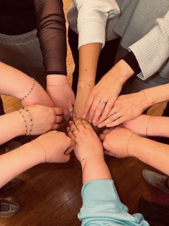 Top-down view of friends' hands joined in a circle, each wearing delicate matching chain bracelets and rings, gathered indoors on a hardwood floor.