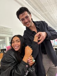 Two smiling friends in a bright industrial-chic studio, a woman in a black leather jacket showing gold bracelets and rings while a man in glasses leans in, white ceiling pipes and clothing racks in the background.