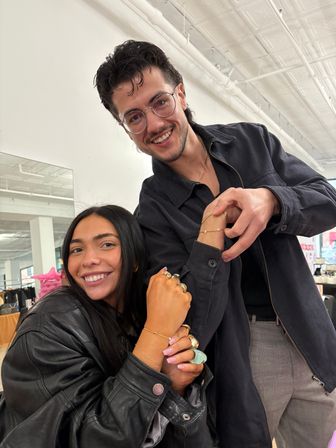 Two smiling friends in a bright industrial-chic studio, a woman in a black leather jacket showing gold bracelets and rings while a man in glasses leans in, white ceiling pipes and clothing racks in the background.