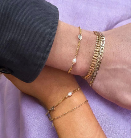 Close-up of two wrists wearing delicate gold chain bracelets with small white pearl accents and a chunky gold chain, styled on a lilac fabric background.