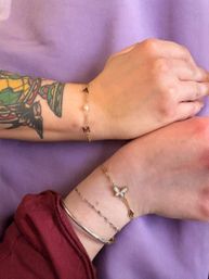 Close-up of two wrists on a purple backdrop wearing stacked delicate gold bracelets — one with a white butterfly charm and arrow, the other with tiny letter and pearl charms; visible colorful forearm tattoo and silver bangle, fashion jewelry detail.