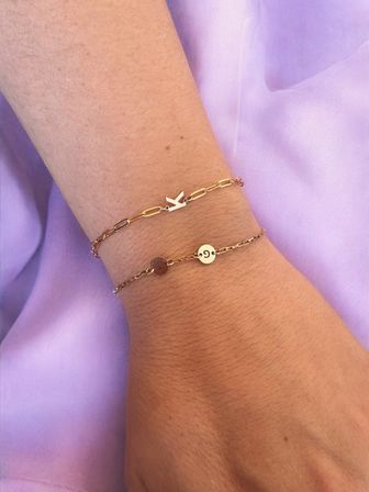 Close-up of a wrist wearing two delicate gold bracelets — a paperclip-style chain with a chevron charm and a thin chain with small round initial discs — on lavender fabric background