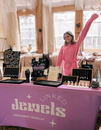 Smiling woman in a pink sweater waves behind a pink table at an indoor artisan market booth showcasing delicate necklaces, rings, and permanent-jewelry displays on black and wooden stands.