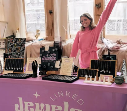 Smiling woman in a pink sweater waves behind a pink table at an indoor artisan market booth showcasing delicate necklaces, rings, and permanent-jewelry displays on black and wooden stands.