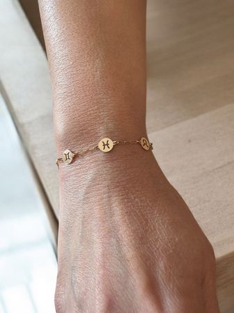 Close-up of a wrist wearing a delicate gold zodiac charm bracelet with small round discs engraved with Gemini, Pisces and Leo symbols, photographed on a light wood surface.