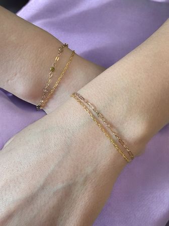 Close-up of two wrists wearing delicate layered gold-tone chain bracelets against a lavender fabric background
