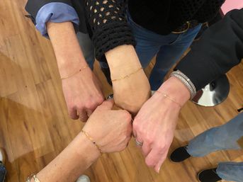 Four wrists joined in a circle displaying delicate gold chain bracelets with tiny gemstones, casual jeans and wooden floor visible — playful indoor shopping moment.