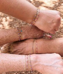 Four diverse wrists stacked, playfully showing layered delicate gold chain bracelets with tiny colorful beads against an outdoor wood-chip ground