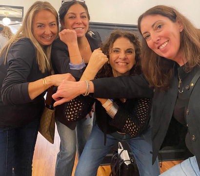 Four smiling women in casual jeans and black tops pose indoors, playfully bumping wrists to show bracelets and bangles.