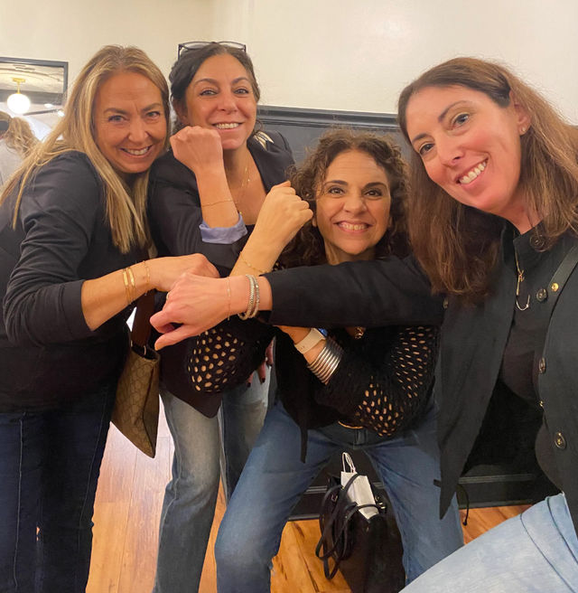 Four smiling women in casual jeans and black tops pose indoors, playfully bumping wrists to show bracelets and bangles.