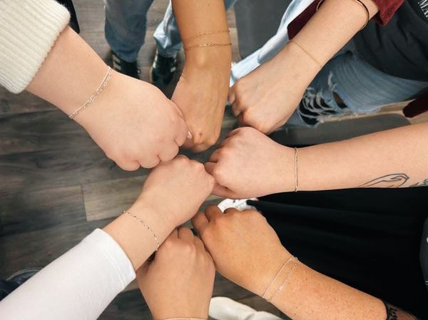 Overhead shot of eight hands joining in a fist-bump circle on a wooden floor, each wrist wearing delicate chain bracelets — casual jeans and a visible tattoo add a playful, team-bonding vibe.