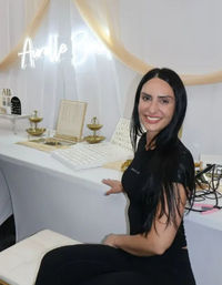 Smiling vendor with long black hair at a white-and-gold jewelry booth displaying ring trays and tiered gold dishes under a glowing neon sign