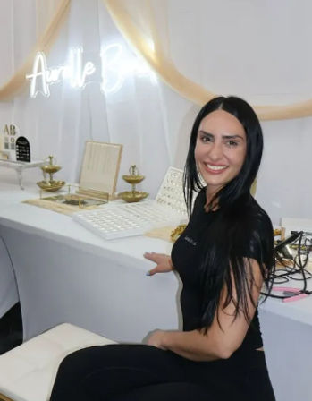 Smiling vendor with long black hair at a white-and-gold jewelry booth displaying ring trays and tiered gold dishes under a glowing neon sign