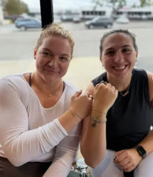 Two smiling friends fist-bumping by a street-facing café window, showing bracelets and casual athleisure with parked cars outside