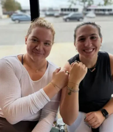 Two smiling friends fist-bumping by a street-facing café window, showing bracelets and casual athleisure with parked cars outside