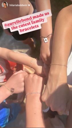 Sunlit close-up of five mixed-age wrists joined in a circle outdoors, each wearing matching delicate chain family bracelets with tiny charms.