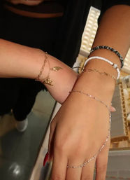 Close-up of two wrists wearing layered bracelets and a delicate hand chain—gold leaf charm, white and black beaded bracelets, and a silver finger chain on a jewelry display.