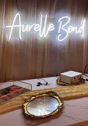 Indoor styling station with glowing cursive neon sign above a table holding an ornate gold mirror, jewelry, tools, and display trays