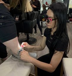 Beauty technician in pink-rimmed magnifying glasses using an LED light to work on a client's ankle above a gray Hoka sneaker in a busy hair salon