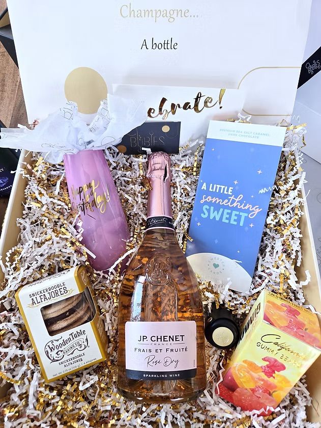 Birthday celebration gift box: sparkling rosé bottle, pink 'Happy Birthday' champagne flute, 'a little something sweet' chocolate bar, snickerdoodle cookies and gummy candies nestled in gold-and-white shredded paper.