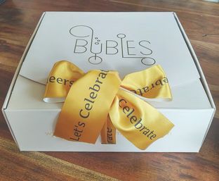 White gift box on a wooden table topped with a shiny gold ribbon bow reading Let's Celebrate, festive party gift packaging
