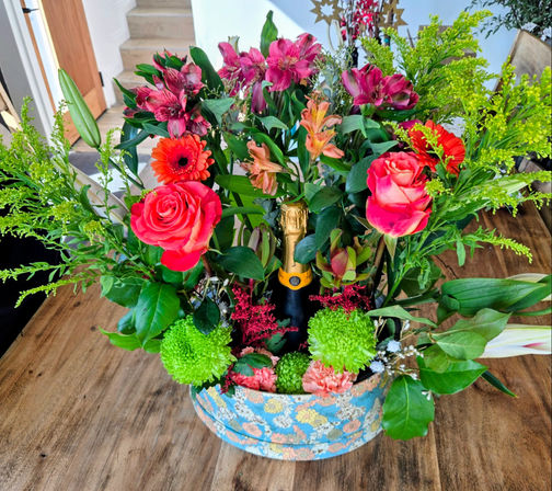 Colorful floral centerpiece on a wooden table featuring pink roses, orange gerbera, green pom‑pom chrysanthemums, alstroemeria and mixed greenery arranged around a champagne bottle in a blue floral round hatbox.